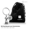A silver meerkat keychain with a round charm and a black velvet pouch are displayed on a white background. Produktbild-Vorschau