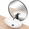 A white oval countertop sink with a black faucet is shown on a wooden surface, with a round mirror reflecting the scene. Náhled obrázku produktu