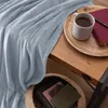 A light blue blanket is draped over a wooden table, next to a mug of tea and a book. Produktbild-Vorschau
