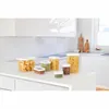 A collection of clear plastic food storage containers filled with various dry goods, arranged on a white countertop in a modern kitchen setting. Voir les images du produit