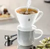 A pour-over coffee maker is shown in use, brewing coffee into a mug. There's also a slice of cake in the background and coffee-related accessories in the foreground. Produktbild-Vorschau