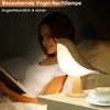 A woman is reading a book in bed at night, with a bird-shaped night lamp and a cup of coffee on the bedside table. Produktbild-Vorschau
