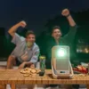 Two young men are watching a movie projected outdoors at night, with snacks and drinks on a table. Produktbild-Vorschau