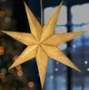 A golden, 3D paper star decoration hangs in front of a blurred background of lights and a window. Náhled obrázku produktu