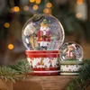 Two Christmas snow globes, one with a nutcracker and the other with a teddy bear, are displayed on a wooden surface with pine branches in the foreground and blurred Christmas lights in the background. Produktbild-Vorschau