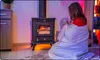 A woman sits on the floor wrapped in a blanket, holding a mug, next to a wood-burning stove with a fire inside. The room is lit with colorful ambient lighting. Voir les images du produit
