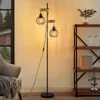 A floor lamp with two cage-style lampshades and wooden accents is placed next to a chair and a bookshelf in a room. Produktbild-Vorschau