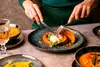 A person is eating a meal with a fork and knife, with several bowls of soup and a stuffed pumpkin on the table. Náhľad obrázku produktu
