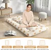 A woman is sitting on a foldable mattress with a bear print. The mattress is on the floor in a room with a wooden floor. Below the mattress, there are diagrams and text indicating different mattress sizes. Produktbild-Vorschau
