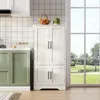 A white kitchen cabinet with four doors stands next to green kitchen cabinets, with a window in the background showing a park scene. Náhľad obrázku produktu