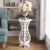 A white decorative stand with a vase of flowers on top, placed next to a bookshelf and a chair. Produktbild-Vorschau