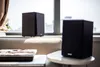 Two black studio speakers are placed on a windowsill and a small table, with a window and curtains in the background. Produktbild-Vorschau