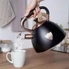 A hand is pouring hot water from a black kettle with copper accents into a white mug. Náhled obrázku produktu