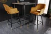 Two mustard yellow velvet bar stools with diamond quilting and gold accents are placed around a dark wood bar table in a modern interior setting. Produktbild-Vorschau