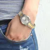 A person's wrist wearing a silver and gold watch, with blue jeans visible. Produktbild-Vorschau
