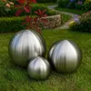 Three metallic spheres of varying sizes are placed on a grassy lawn in a garden setting. A winding path and flowering plants are visible in the background. Produktbild-Vorschau