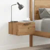 A close-up view of a wooden bedside table with a drawer, a lamp, and a cup of coffee on top, next to a bed. Náhled obrázku produktu