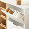 A close-up view of a white shoe cabinet with its top flap open, revealing shoes inside. Another section of the cabinet with wooden shelves is visible to the left. Náhľad obrázku produktu