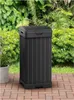 A black trash can sits on a brick patio surrounded by lush green foliage. Anteprima dell’immagine prodotto