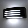 A black modern outdoor wall lamp mounted on a light gray wall, emitting a bright white light downwards and through its horizontal slats. Anteprima dell’immagine prodotto