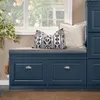 A blue shoe cabinet with a gray cushion and decorative pillows sits in front of a window with greenery outside. Produktbild-Vorschau