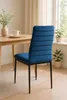 A blue velvet dining chair is shown from the back and side, placed next to a wooden dining table with a vase of greenery and a mug on it. Plants are visible in the background near a window. Produktbild-Vorschau