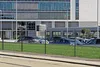 A fence in front of a modern office building with cars parked in the background. Produktbild-Vorschau