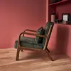 A green velvet armchair with wooden legs is placed in a room with pink walls and a wooden floor. Shelves with decorative items are visible in the background. Produktbild-Vorschau