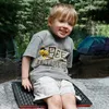 A young boy is sitting on a mat outdoors, with tents and trees in the background. Produktbild-Vorschau