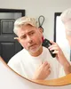 A man is shaving his face with an electric razor in front of a mirror. Voir les images du produit