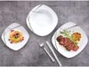 A set of square white plates with a modern abstract design, featuring a stack of pancakes on one plate, a sliced steak with rosemary on another, and a large soup bowl. Cutlery is also visible. Náhled obrázku produktu