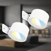Two modern white wall lights are shown in a dimly lit room with a blurred background of furniture and a kitchen area. Anteprima dell’immagine prodotto