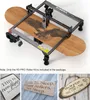 A laser engraver is shown on a wooden board with an eagle design, accompanied by smaller images of engraved items. Náhled obrázku produktu