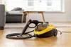 A yellow and black steam cleaner with its hose and nozzle is placed on a wooden floor in a living room setting. Náhled obrázku produktu