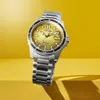 A close-up studio shot of a Citizen Eco-Drive watch with a gold dial and silver band, set against a bright yellow background with subtle shadows. Produktbild-Vorschau