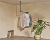 A corner of a room with a ceiling-mounted clothing rack displaying two shirts and a jacket, a wooden stool, and a potted plant. Produktbild-Vorschau