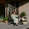 A woman sits in a hanging egg chair on a wooden deck, with plants and flowers around her. Produktbild-Vorschau