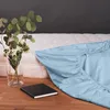 A light blue fitted sheet is draped over a mattress, with a vase of flowers and a notebook on a wooden surface in the foreground. Náhled obrázku produktu