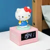 A pink digital alarm clock with a Hello Kitty figure on top, placed on a white surface next to a wooden bed frame. Podgląd zdjęcia produktu