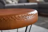 Close-up shot of a round leather ottoman with decorative stitching, resting on thin black legs. A blurred living room background with a grey sofa is visible. Produktbild-Vorschau