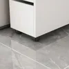 A close-up view of the bottom of a white cabinet with black wheels on a gray tiled floor. Produktbild-Vorschau