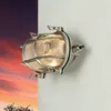 A wall-mounted outdoor lamp with a ribbed glass shade and a metal cage, set against a dramatic sunset sky on the left and a plain white wall on the right. Produktbild-Vorschau