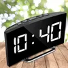 A digital alarm clock with a mirrored LED display sits on a wooden surface, with a blurred green background of foliage. Produktbild-Vorschau