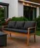 A wooden garden bench with black cushions is placed on a wooden deck, with lush green bushes and flowers in the background. Produktbild-Vorschau