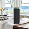 A black Tronsmart T8 outdoor speaker is placed on a wooden table with a white cloth in front of it, in a kitchen setting with a stove and windows in the background. Náhľad obrázku produktu