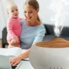 A woman is working on a laptop while holding a baby, with an aroma diffuser in the foreground emitting steam. Podgląd zdjęcia produktu