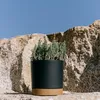 A black planter with a cork base sits on a rocky surface in front of large rocks and a blue sky. Náhľad obrázku produktu