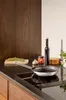 A stainless steel frying pan is placed on a black induction hob, with ingredients like herbs, garlic, and a jar of tomatoes nearby, suggesting preparation for cooking. Podgląd zdjęcia produktu
