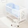 A white crib with blue bedding and a teddy bear sitting on a white rug in front of it. Voir les images du produit