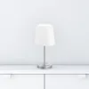 A white fabric lampshade table lamp with a silver base and stem sits on a white surface against a white wall. Podgląd zdjęcia produktu